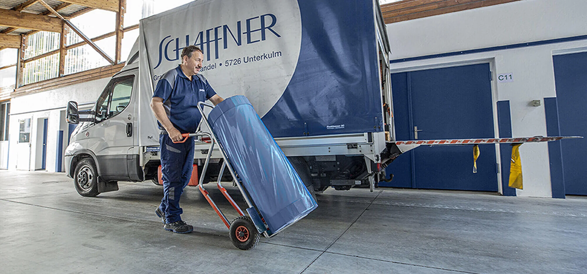 Schaffner GF AG Mitarbeiter schiebt beladenen Transportwagen in Lagerhalle vor firmeneigenem Lieferwagen - zuverlässiger Direktlieferservice für die grafische Industrie in der Schweiz